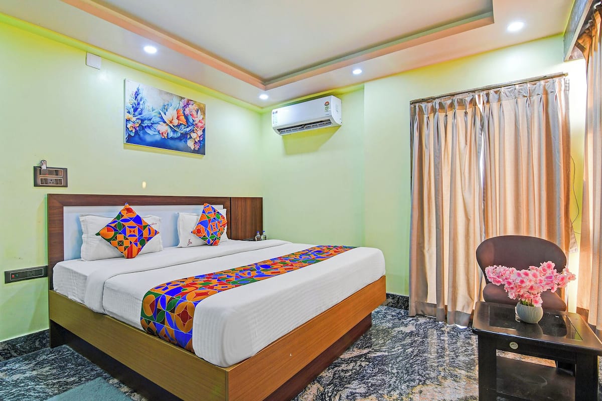 A hotel room with a bed, a painting, an air conditioner, a curtain, and a vase with flowers.