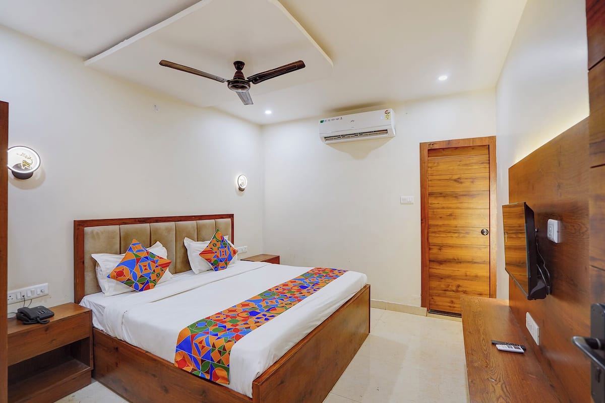 A bedroom with a bed, a ceiling fan, an air conditioning unit, a wooden door, and a bedside table with a phone.