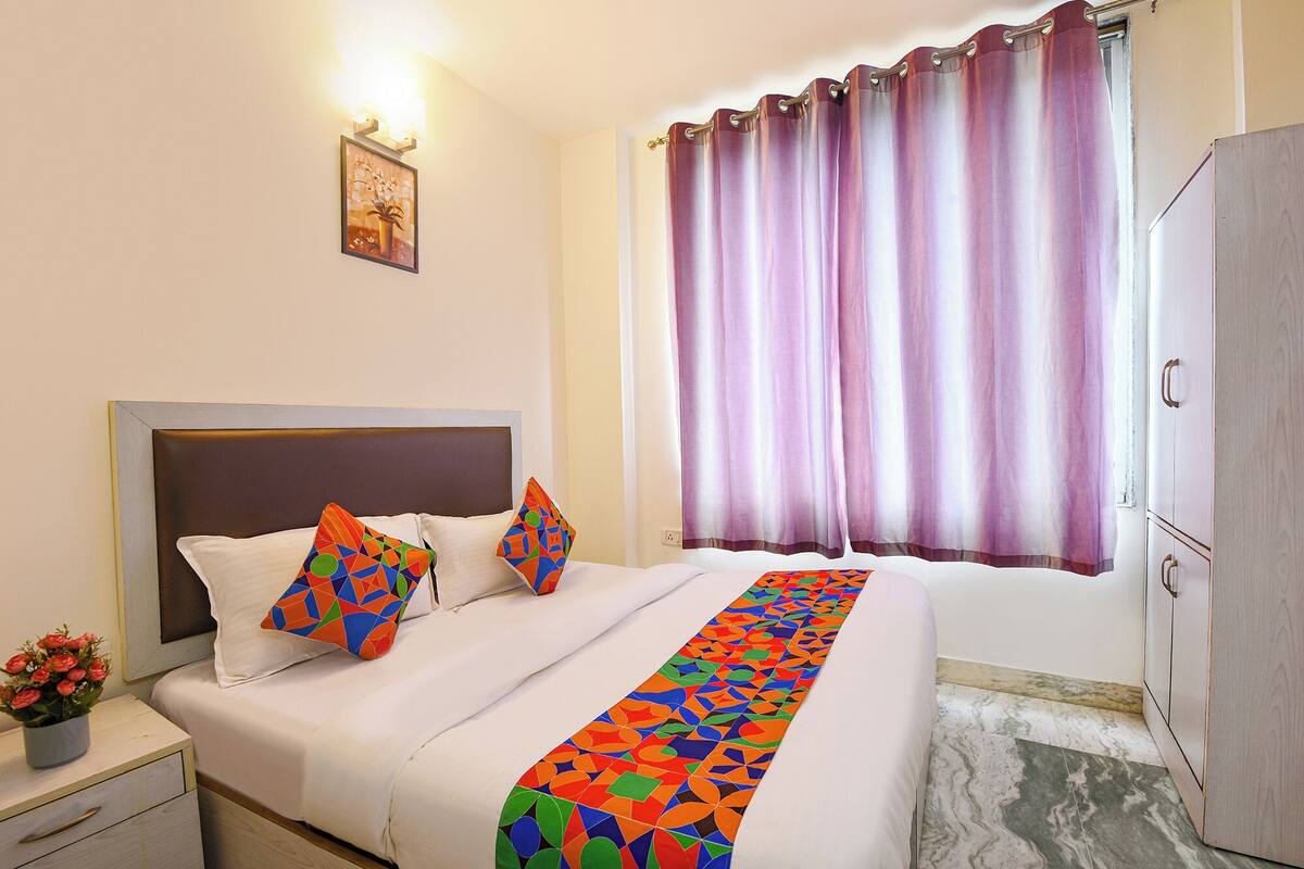 Ein Hotelzimmer mit Bett, Nachttisch mit Blumenvase, Kleiderschrank und Fenster mit violetten Vorhängen.