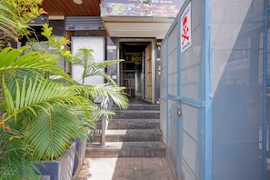 Um corredor exterior estreito com degraus que levam à entrada de um edifício, rodeado por vegetação.