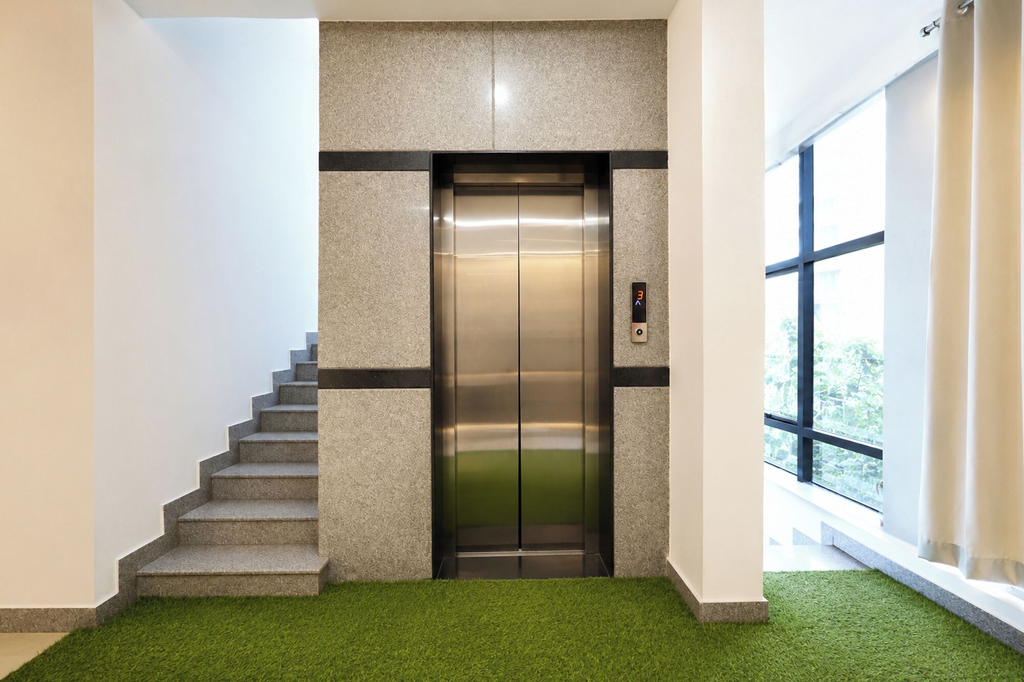 An elevator in a building with a staircase leading to it, a digital display, and a window with a view of greenery outside.