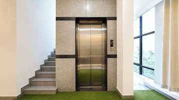 An elevator in a building with a staircase leading to it, a digital display, and a window with a view of greenery outside.