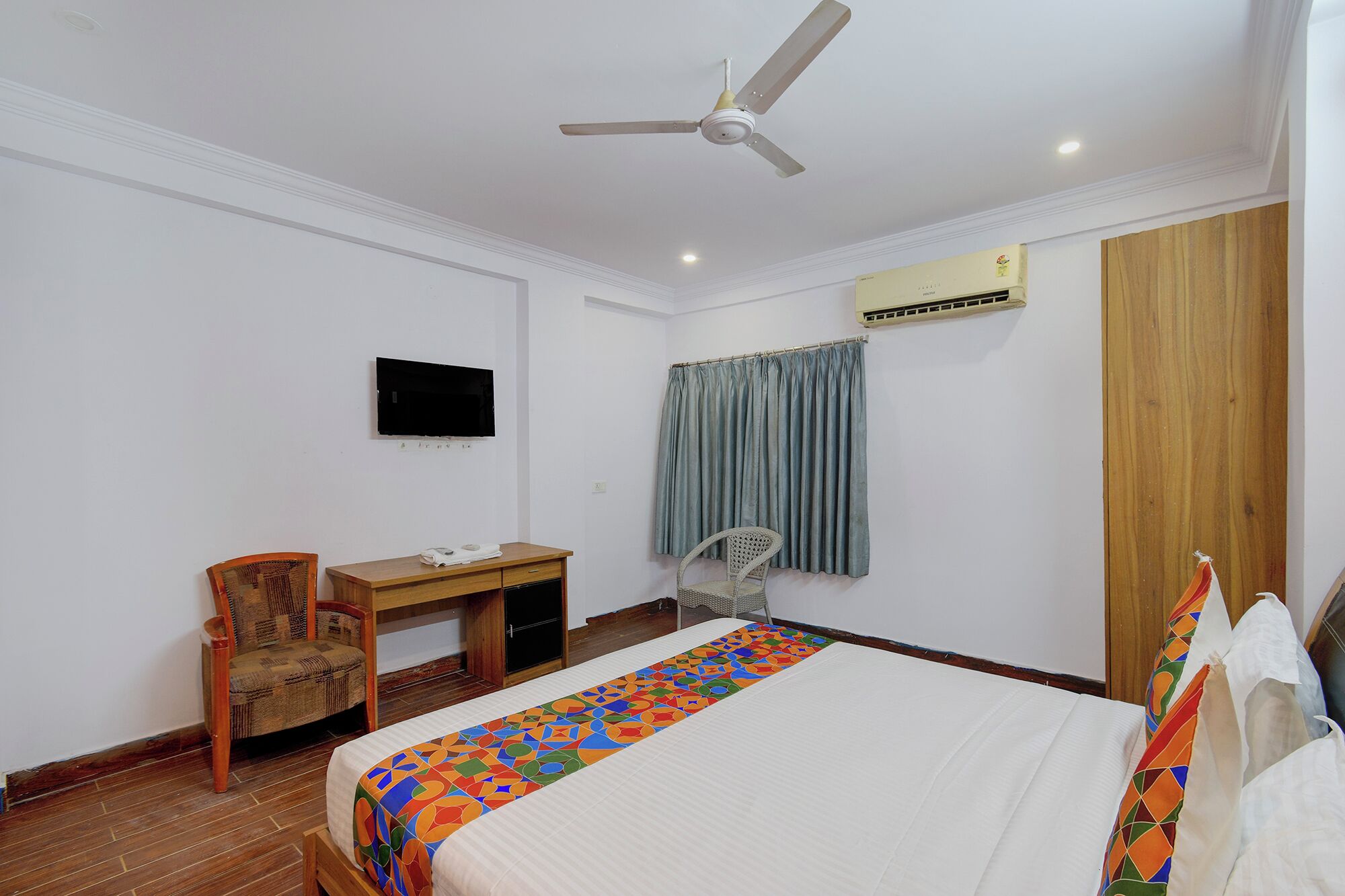 Habitación de hotel con cama, escritorio, silla, ventilador de techo, unidad de aire acondicionado y televisor montado en la pared.