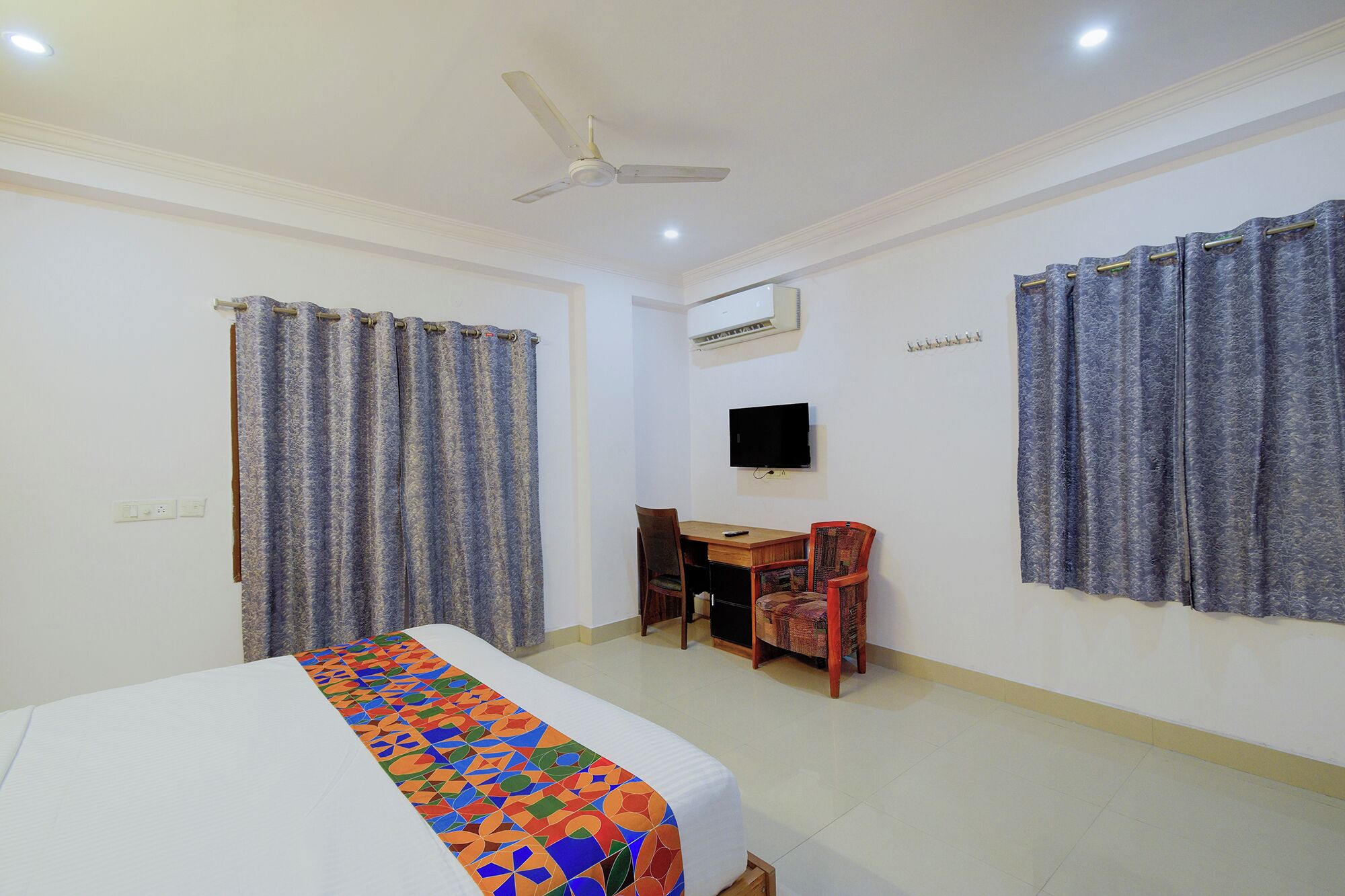 Habitación de hotel con cama, escritorio, silla, ventilador de techo y televisor.