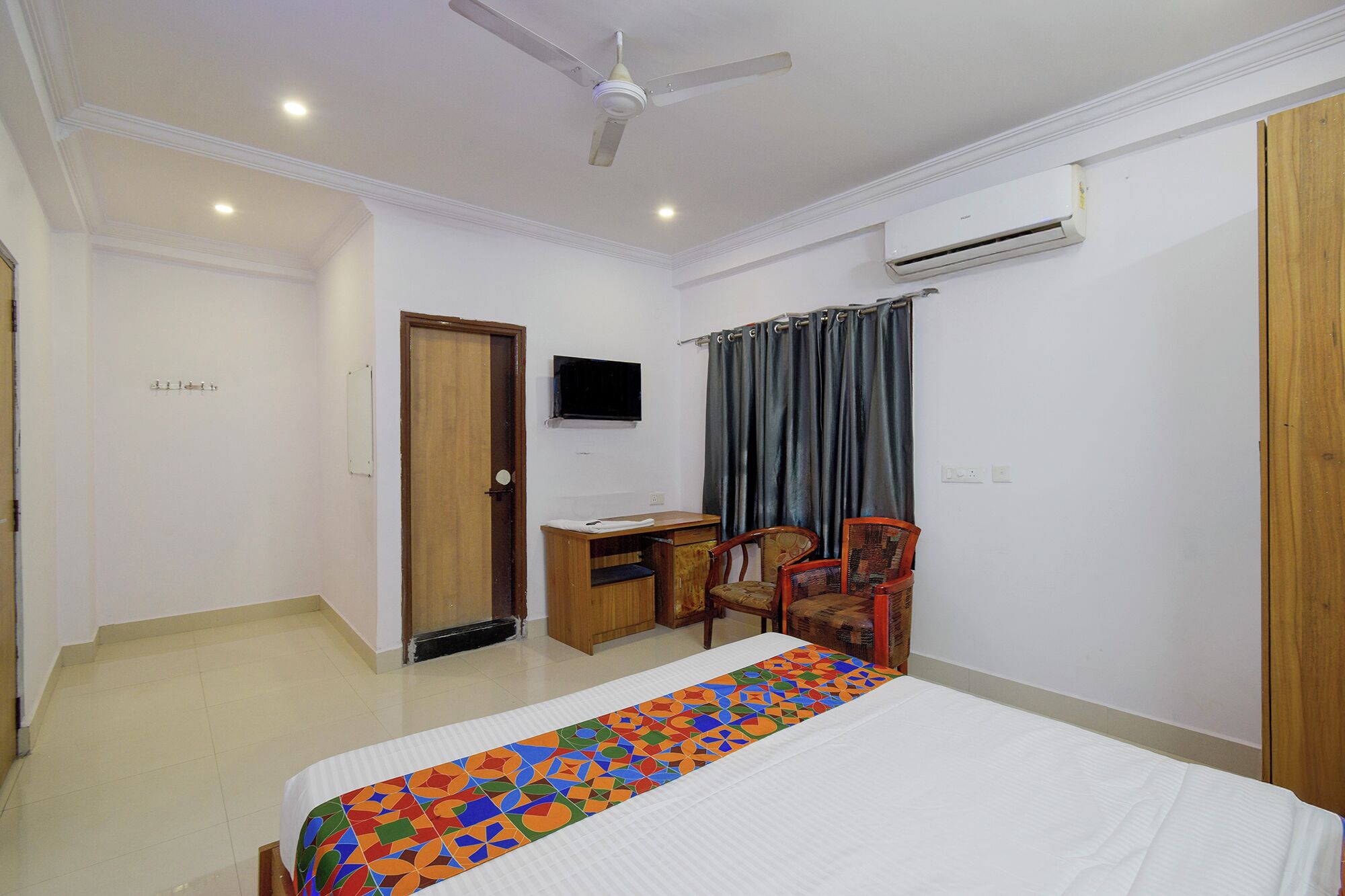 A hotel room with a bed, desk, two chairs, a ceiling fan, an air conditioning unit, and a TV.