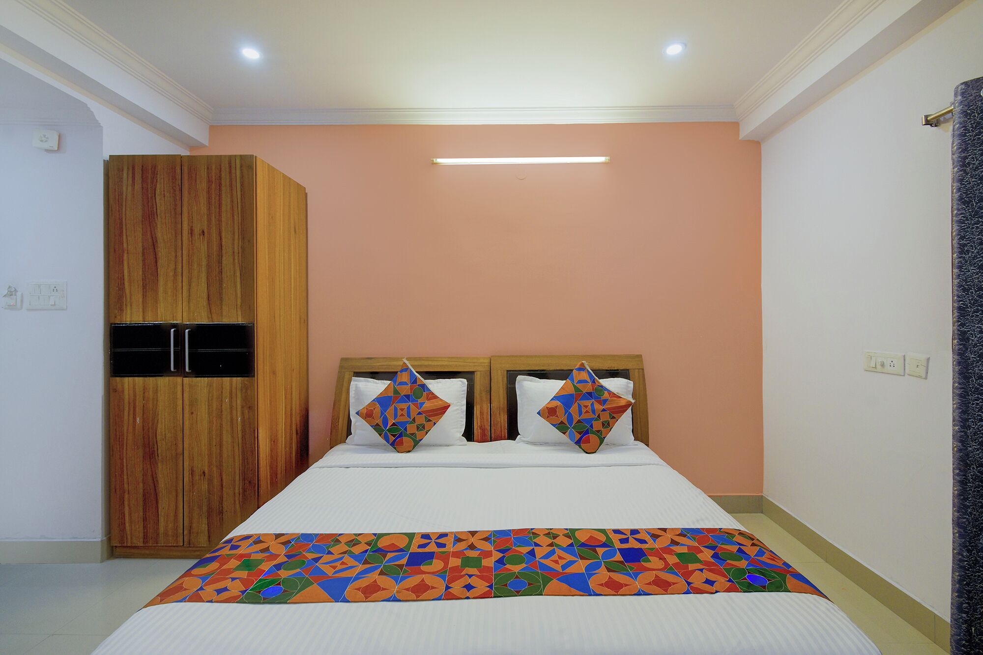 A bedroom with a bed, two pillows with geometric patterns, a wooden wardrobe, and a ceiling light.