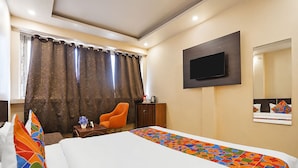 A hotel room with a bed, a TV mounted on the wall, a chair, and a small table with a vase of flowers. - Fahotel Rudransh Residency-1Km from Kashi Vishwanath Temple (Varanasi)