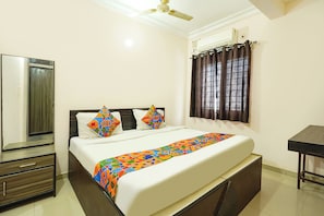 A bedroom with a bed, a bedside table, a wardrobe, a window with curtains, and a ceiling fan. - Fabhotel Utsav Palace Inn (Bangalore)