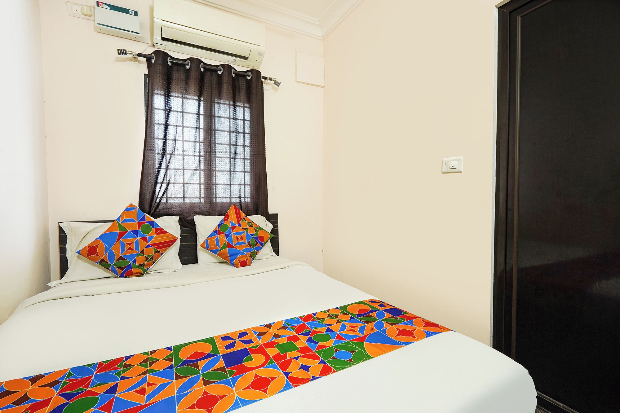 A bed with a colorful geometric-patterned bedspread, a window with a dark curtain, and an air conditioning unit on the wall.