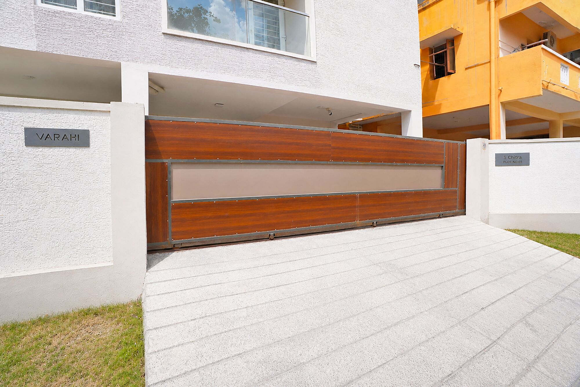 A modern building with a wooden gate, a nameplate reading 'VARAHI,' and a sign indicating 'S Chira Plot No. 65.'