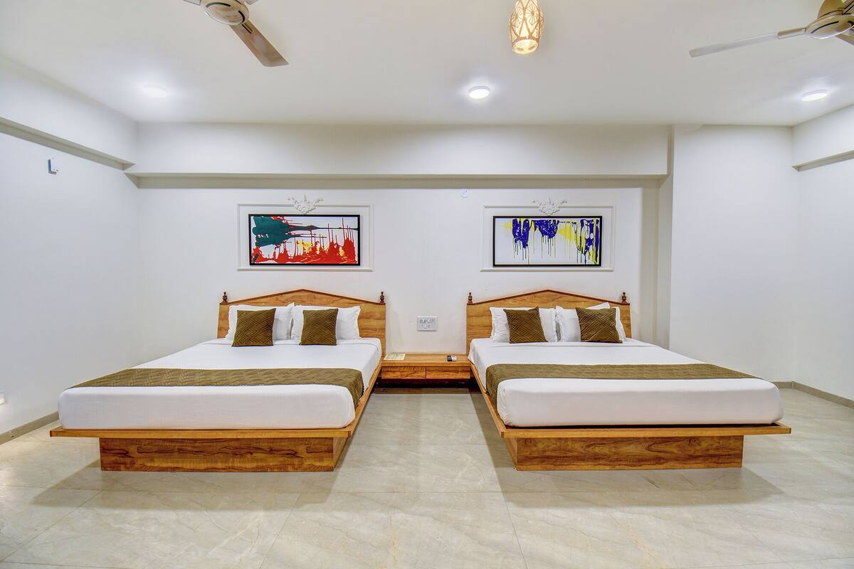 Quarto de hotel com duas camas de madeira, um ventilador de teto, iluminação embutida e duas pinturas abstratas nas paredes.