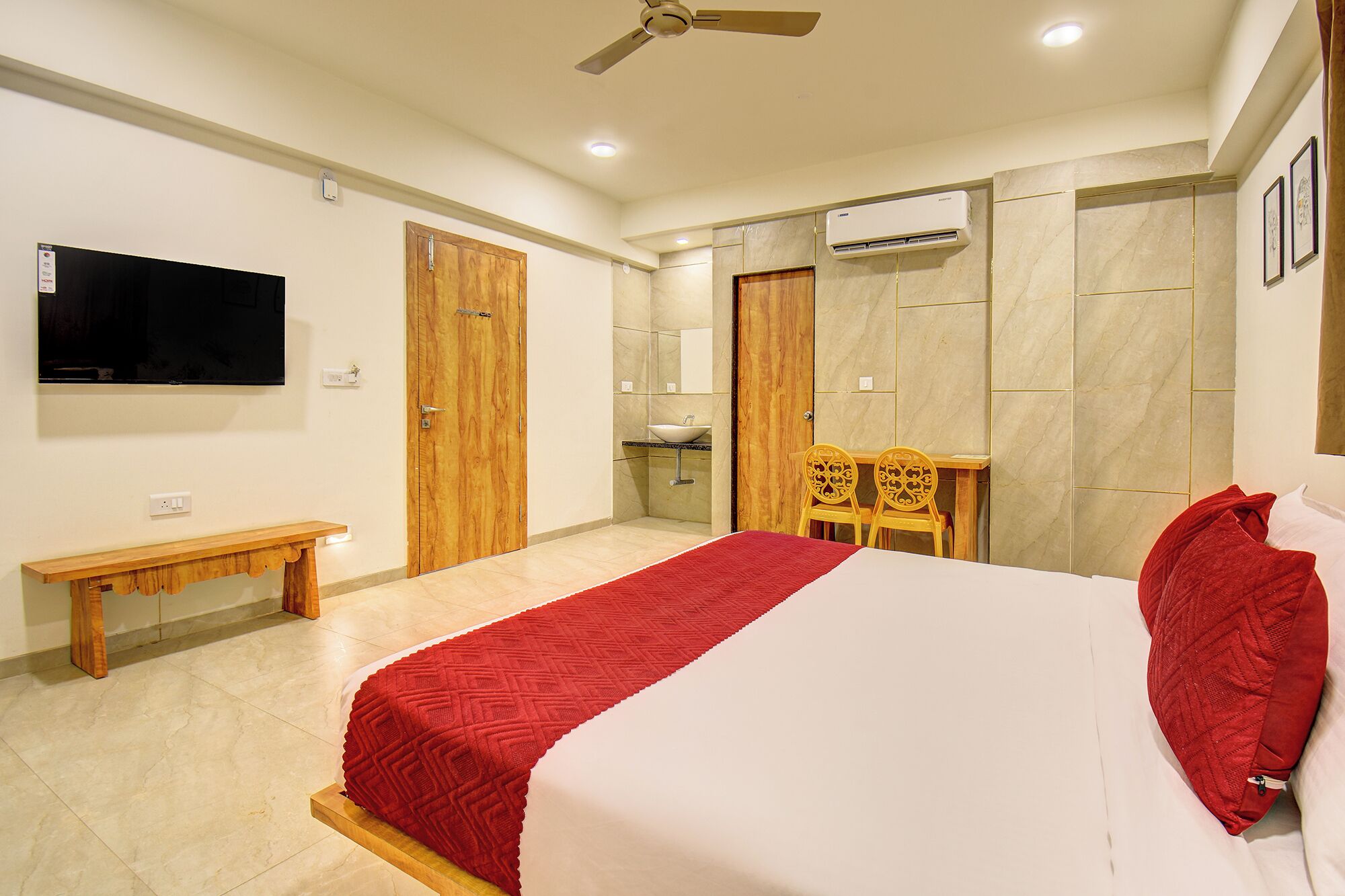 A bedroom with a bed, a bench, a TV, a ceiling fan, an air conditioning unit, and a bathroom with a sink and mirror.