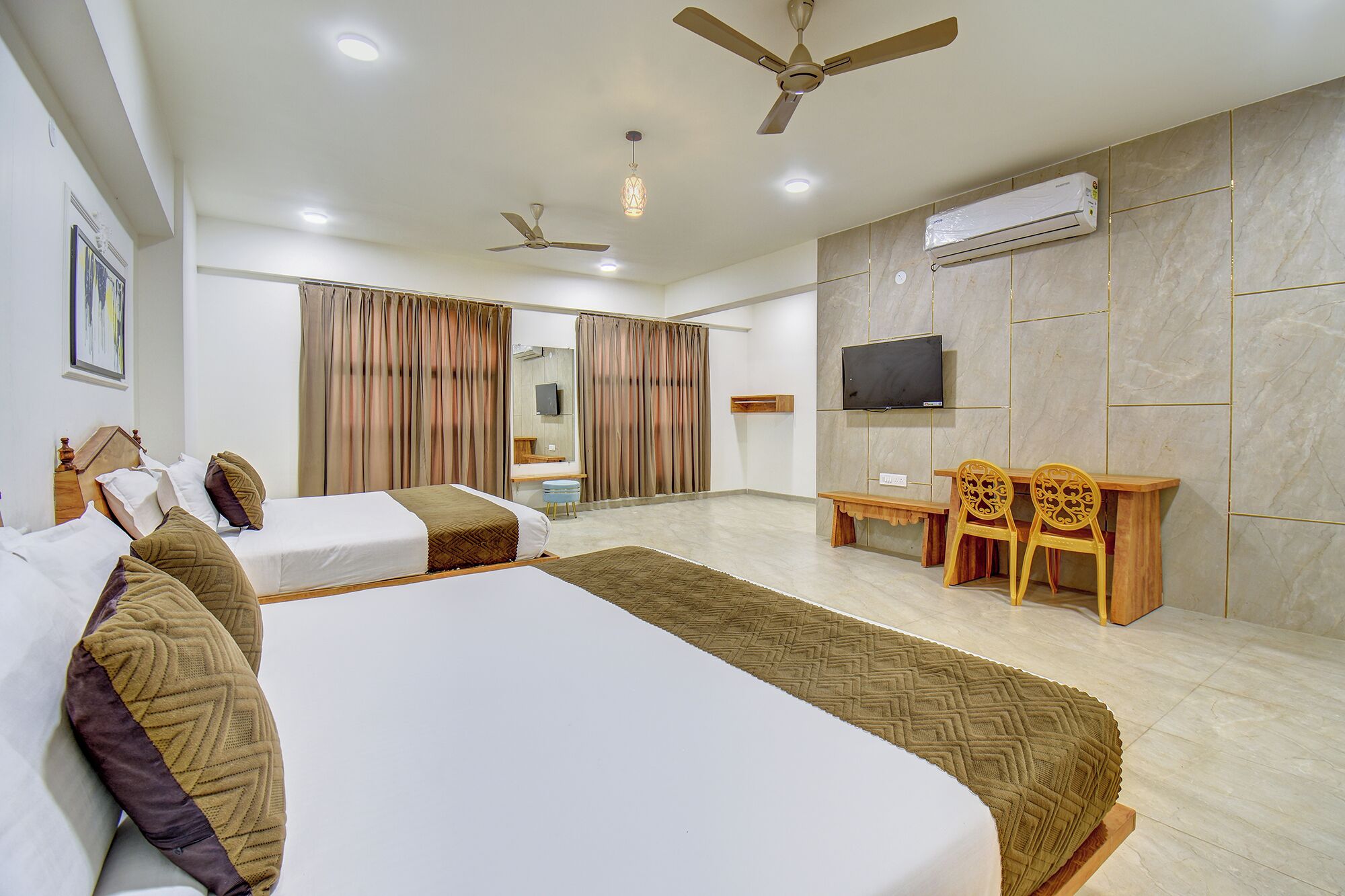 Habitación de hotel con dos camas, televisión, ventiladores de techo y un equipo de aire acondicionado, junto a una pequeña mesa con sillas.