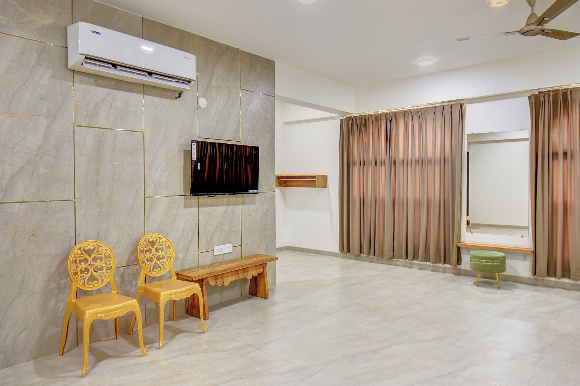 Una sala de estar moderna con un televisor de pantalla plana, equipo de aire acondicionado, dos sillas amarillas, un banco de madera y un reposapiés verde.