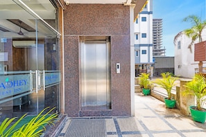 Entrada de hotel com porta de vidro, elevador de metal e plantas em vasos do lado de fora.