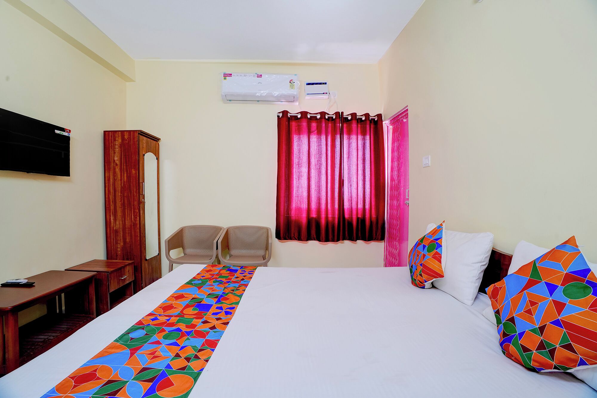 A hotel room with a bed, a desk, two chairs, a TV, and a window with curtains.