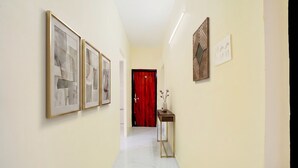 A hallway with framed abstract art, a red door, a small table with a vase, and a wall-mounted light fixture.