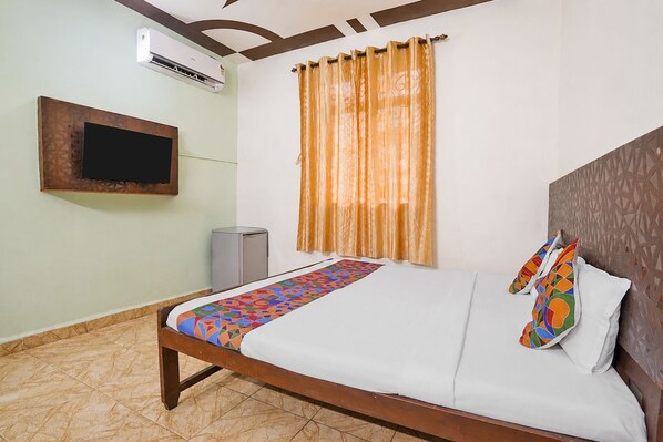 A hotel room with a bed, a television mounted on the wall, an air conditioning unit, a window with curtains, and a patterned floor.