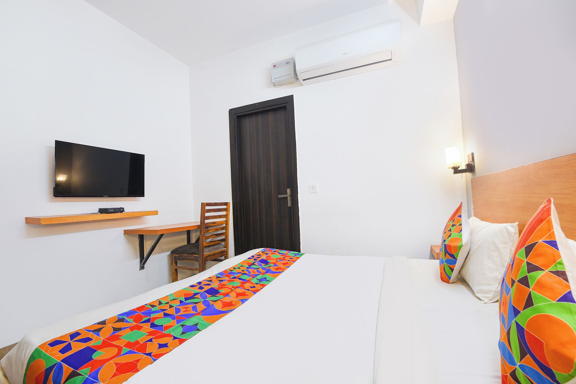 A hotel room with a bed, a TV, a desk, and an air conditioning unit.