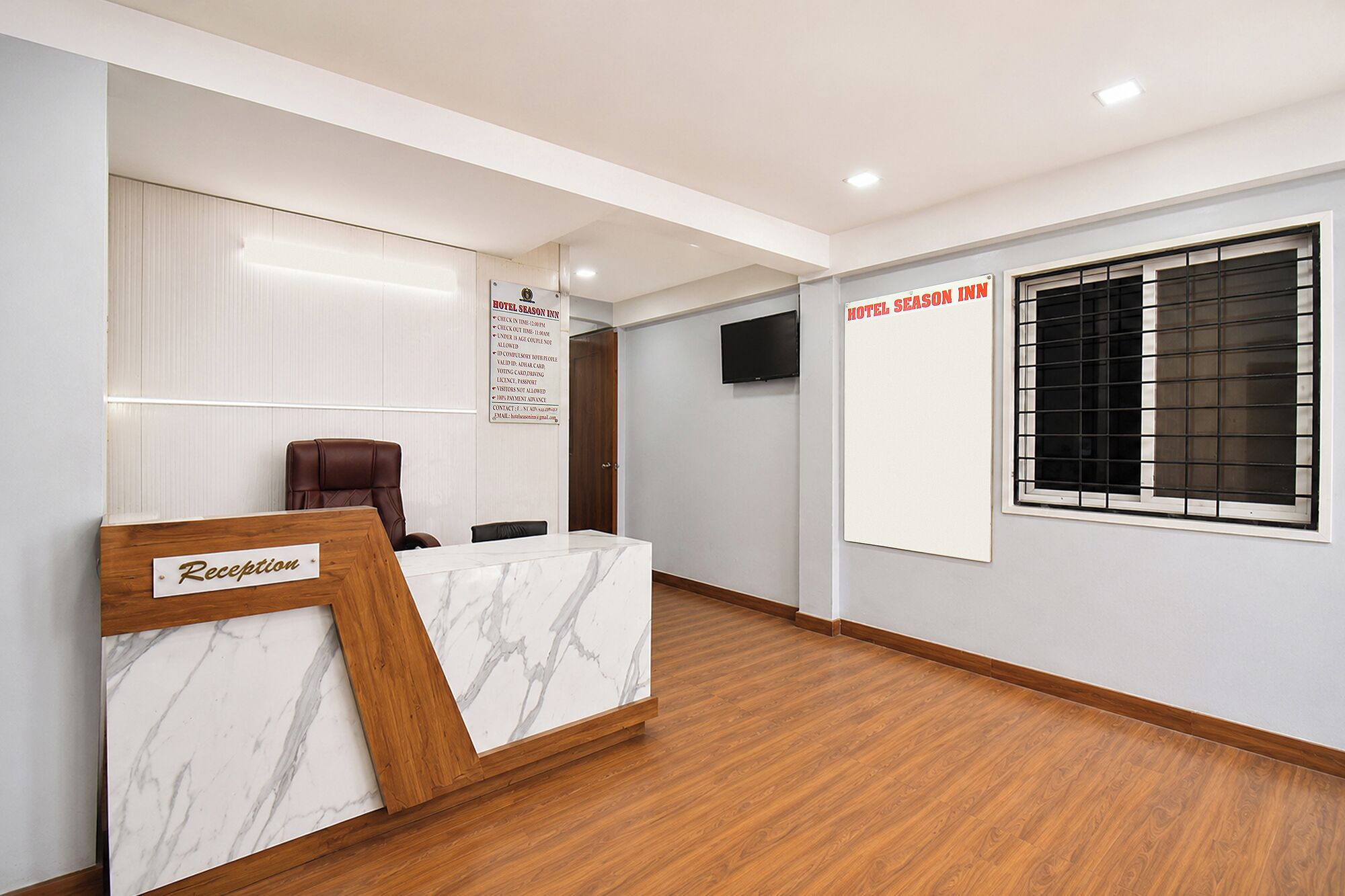 Ein Hotel-Rezeptionsbereich mit einem Marmor-Empfangstresen, einer Holzeinbauscheibe, einem an der Wand befestigten Fernseher und einem Fenster mit Metallgitter.