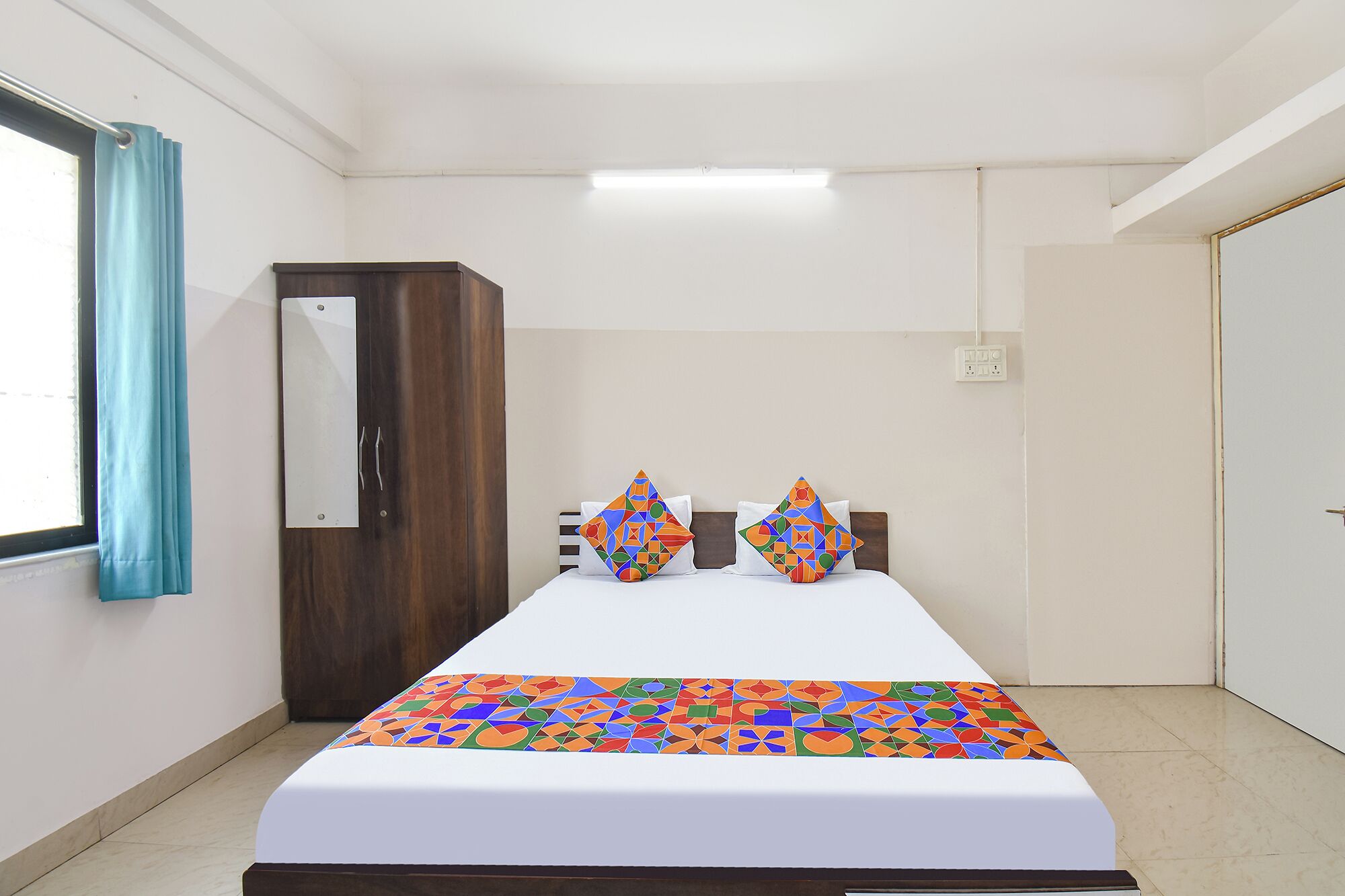 A hotel room with a bed, two pillows, a wooden wardrobe, a window with curtains, and a door.