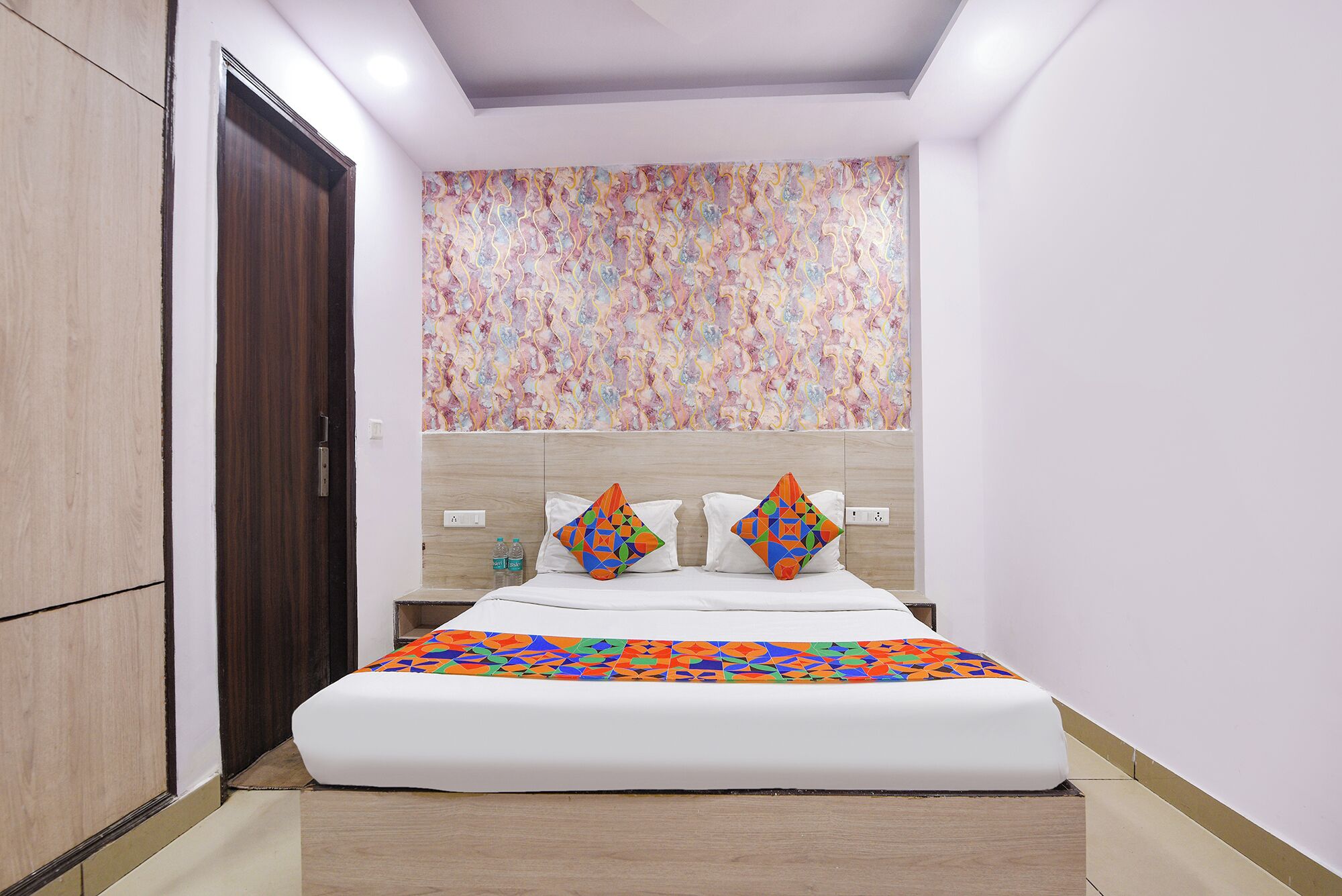 A hotel room with a bed, two pillows, a nightstand, and a wall with a colorful abstract pattern.