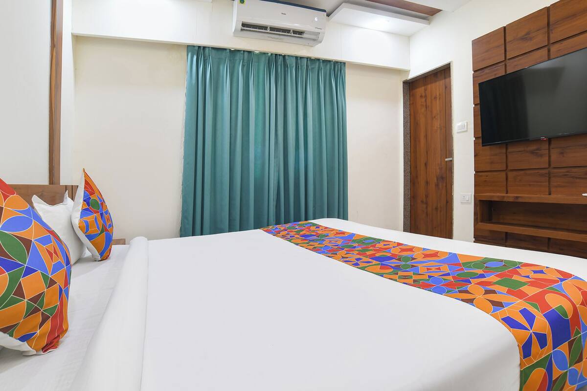 Una habitación de hotel con una cama, un televisor montado en un panel de madera, una ventana con cortinas y un equipo de aire acondicionado.