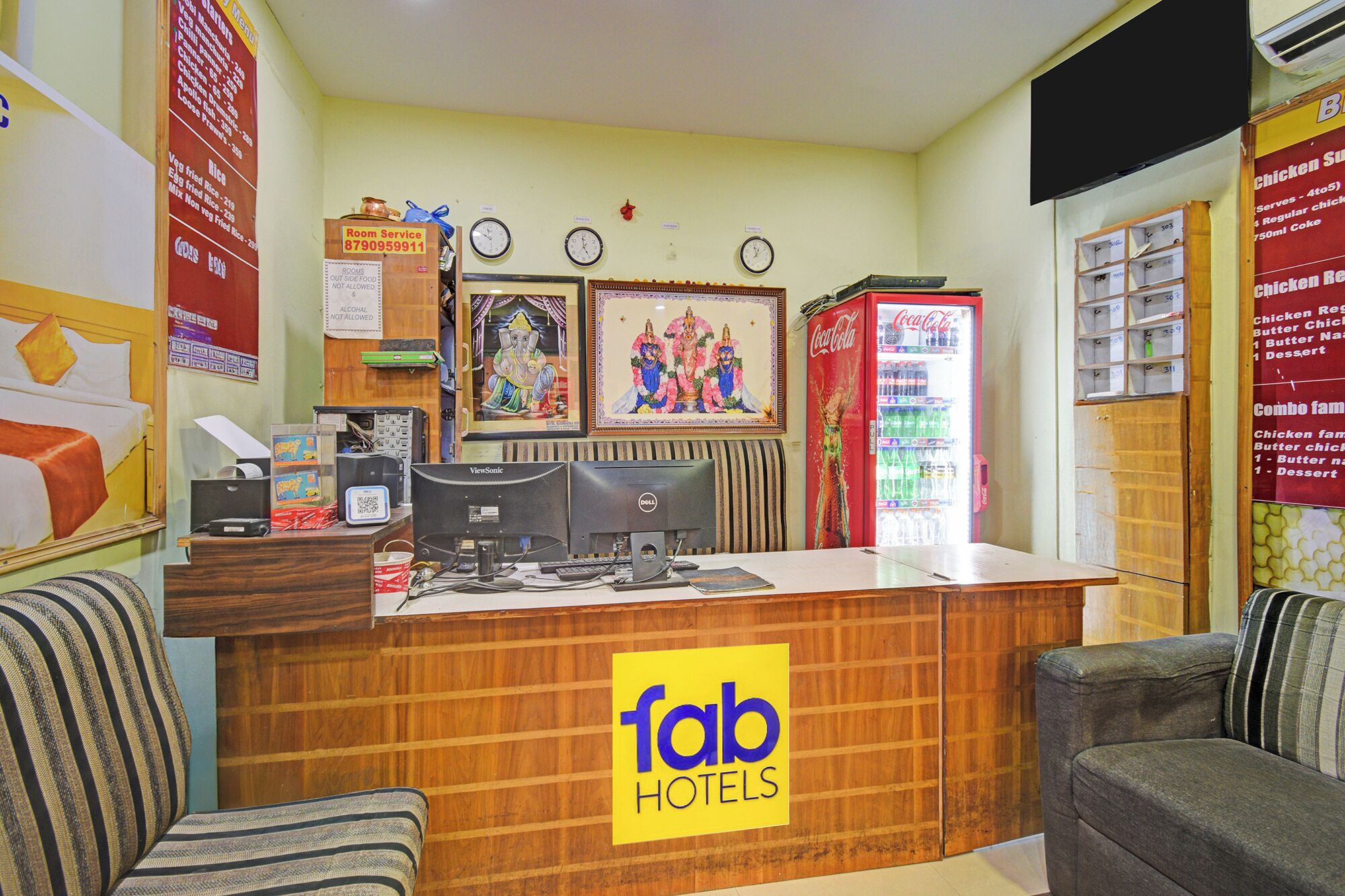 Un espace d’accueil d’hôtel avec un comptoir en bois, deux horloges, un réfrigérateur Coca-Cola et un panneau indiquant « Fab Hotels ».