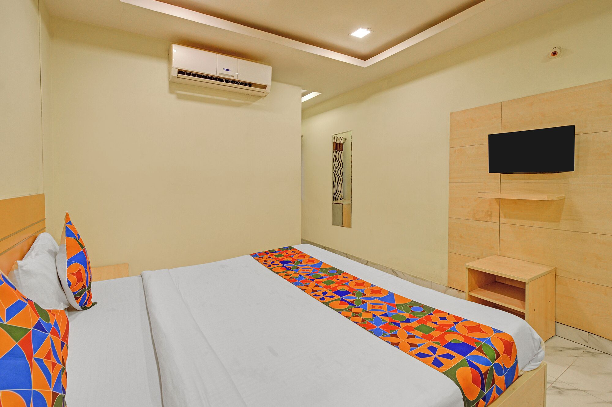 A hotel room with a bed, an air conditioning unit, a television mounted on the wall, and a small shelf.