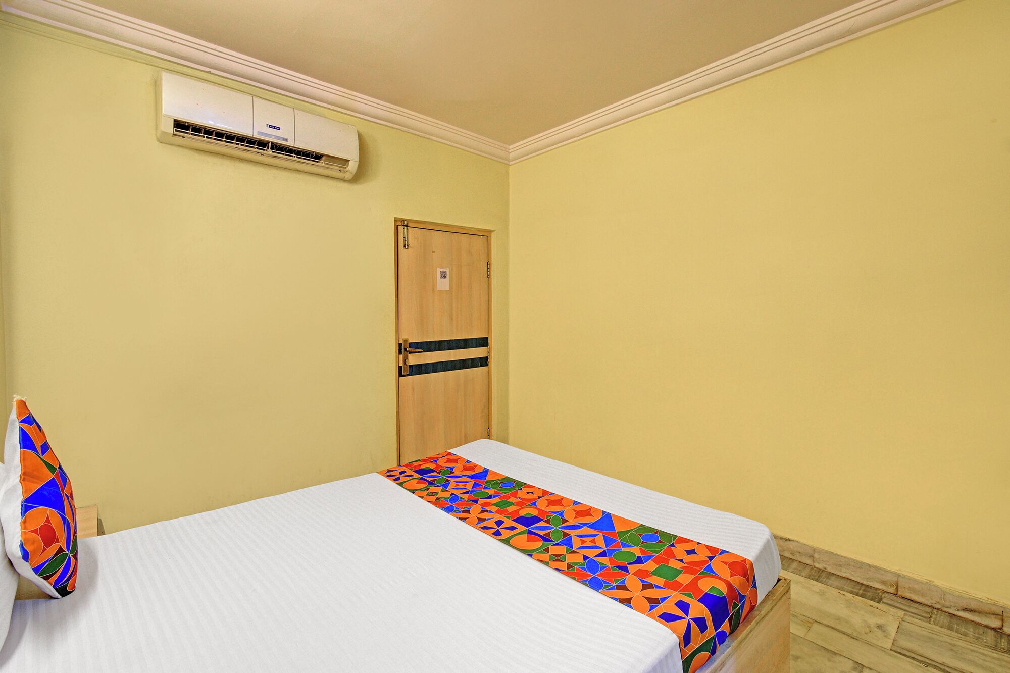 A small room with a bed, an air conditioning unit, a wooden door, and a colorful pillow.