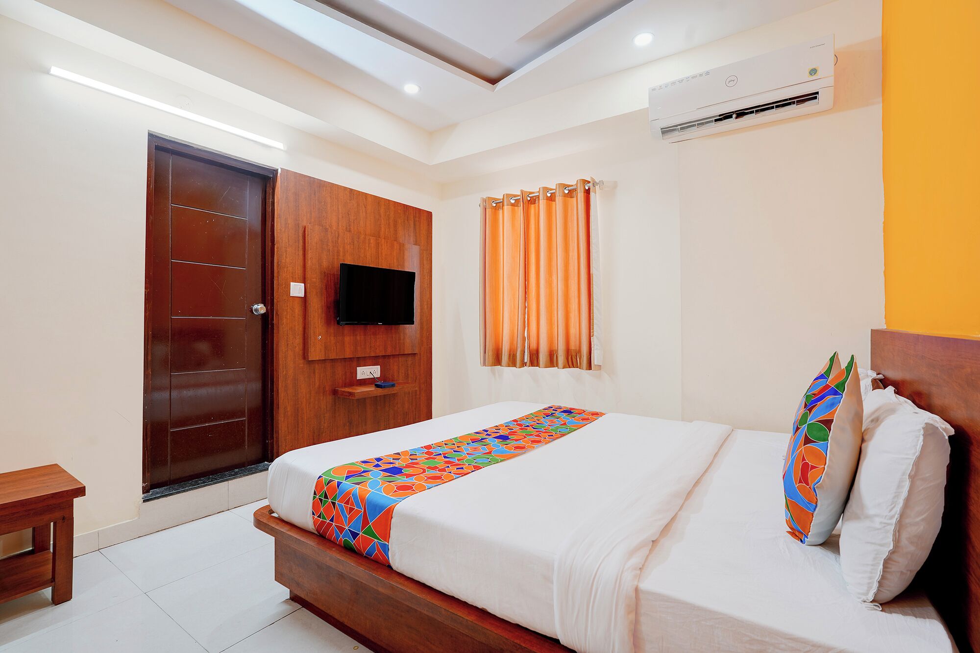 A hotel room with a bed, a television mounted on the wall, an air conditioning unit, a wooden door, and a window with curtains.