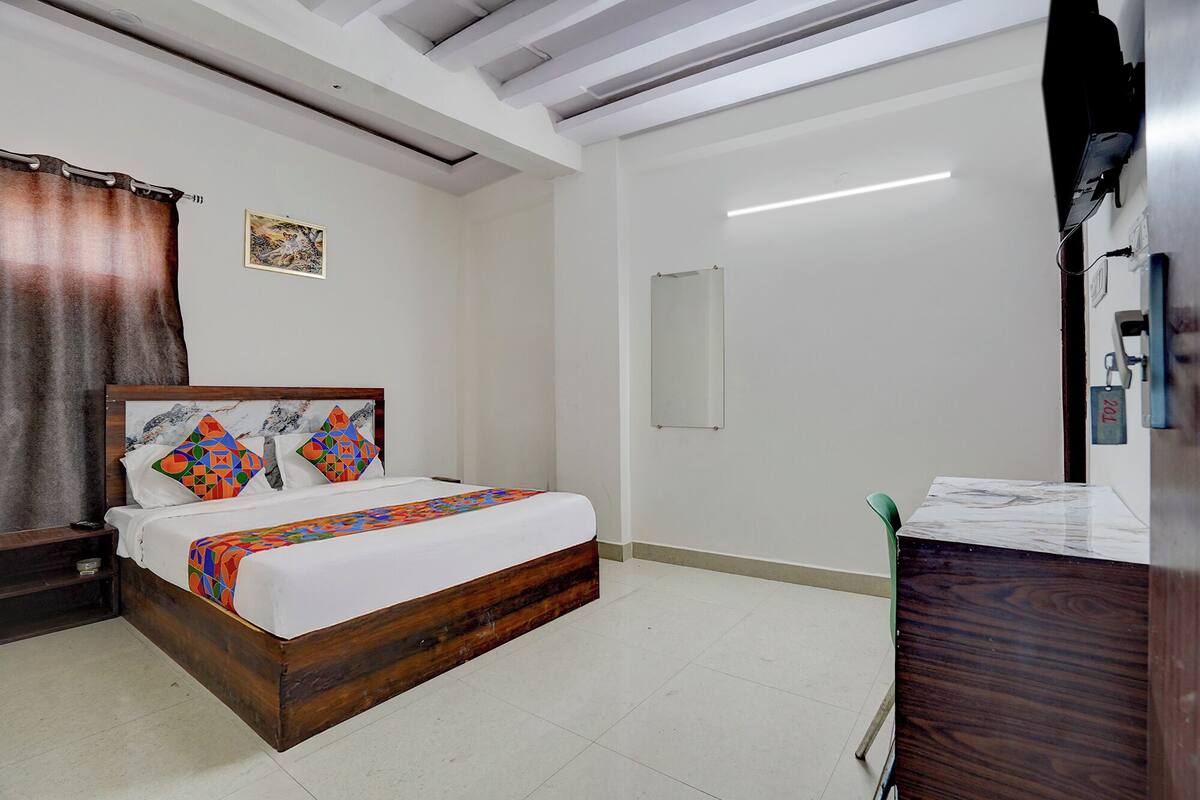 Habitació d'hotel amb un llit de fusta, un escriptori petit, una cadira, un mirall i una paret blanca.