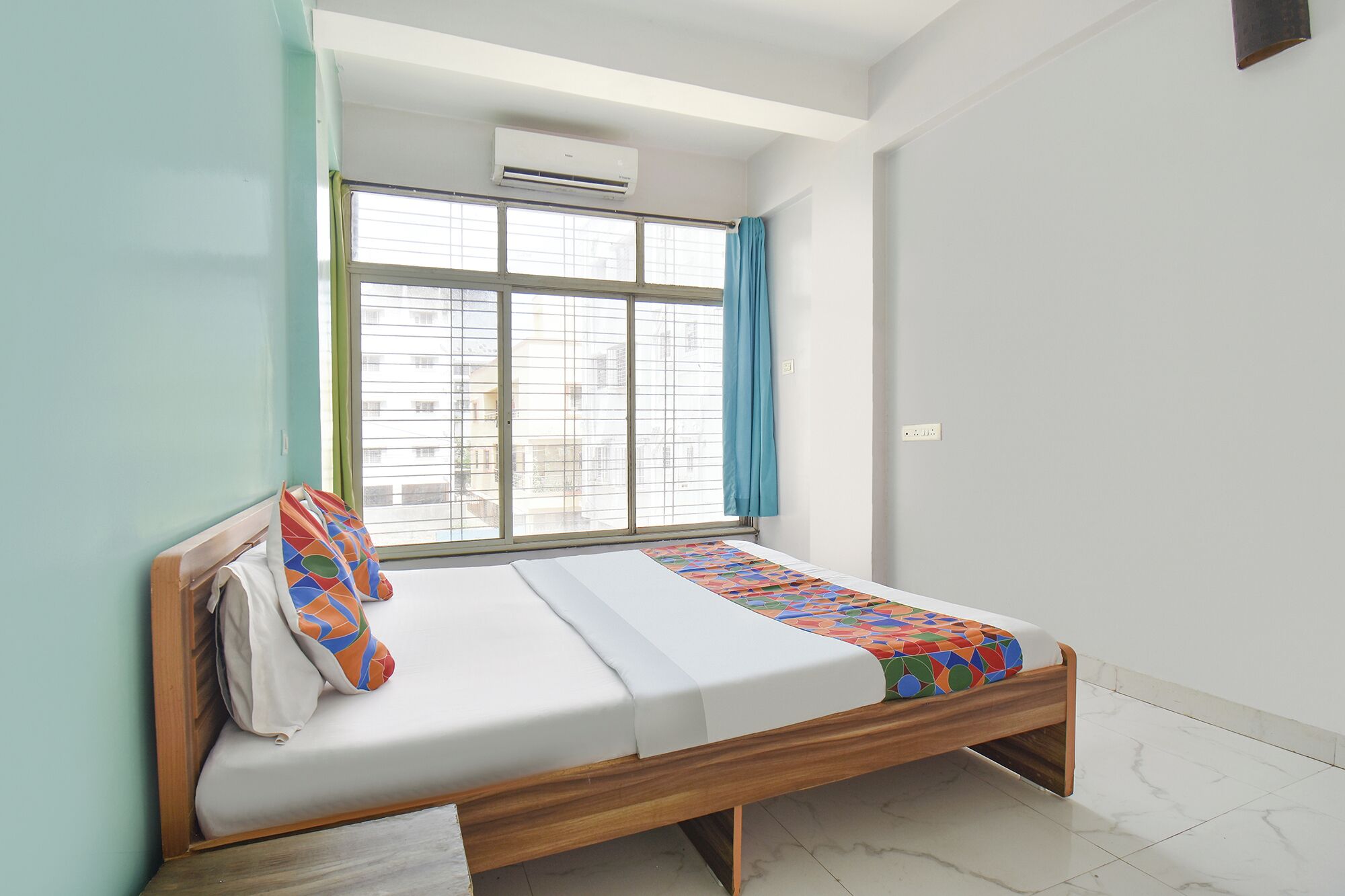 A single bed with a wooden frame, a window with a view of buildings, and an air conditioning unit above the window.