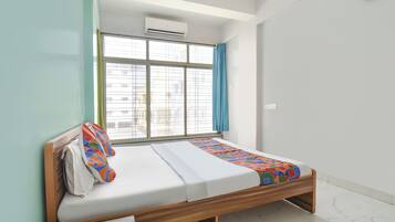 A single bed with a wooden frame, a window with a view of buildings, and an air conditioning unit above the window.