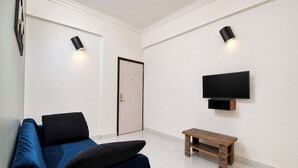 A room with a blue sofa, a small wooden bench, a TV mounted on the wall, and two wall-mounted lights. - Via Dazling Studio (Pune)