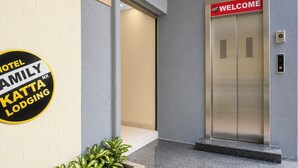 L’entrée d’un hôtel, avec une pancarte « BIENVENUE » au-dessus d’un ascenseur, une autre pancarte indiquant « HÔTEL FAMILY KATTA LODGING » et une plante en pot.
