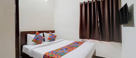 Une chambre d’hôtel comprenant un lit, des oreillers, une couvre-lit à motifs, une fenêtre avec des rideaux, un appareil de climatisation et un téléviseur à écran plat fixé au mur.
