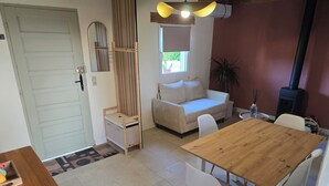 Una habitación acogedora con una mesa de comedor de madera, sillas blancas, un sofá, un pequeño banco de madera, un espejo y una ventana con vistas a zonas verdes.