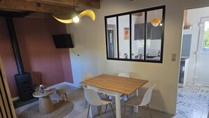Un comedor con una mesa de madera y sillas blancas, un estufa de leña negra, un televisor en la pared y un ventanal con vistas al exterior.