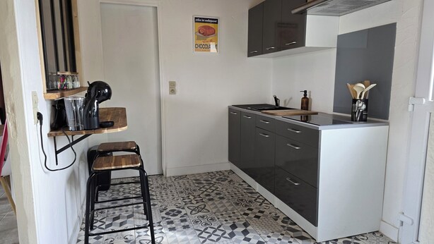 Cocina compacta con suelo de baldosas estampadas, un taburete pequeño, cafetera y lámpara colgante.
