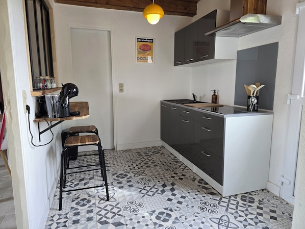 A compact kitchen with a patterned tile floor, a small stool, a coffee maker, and a hanging light fixture.