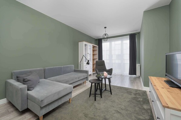 Een woonkamer met een grijze bank, een klein rond tafeltje met een stoel, een wit kastje en een televisie.