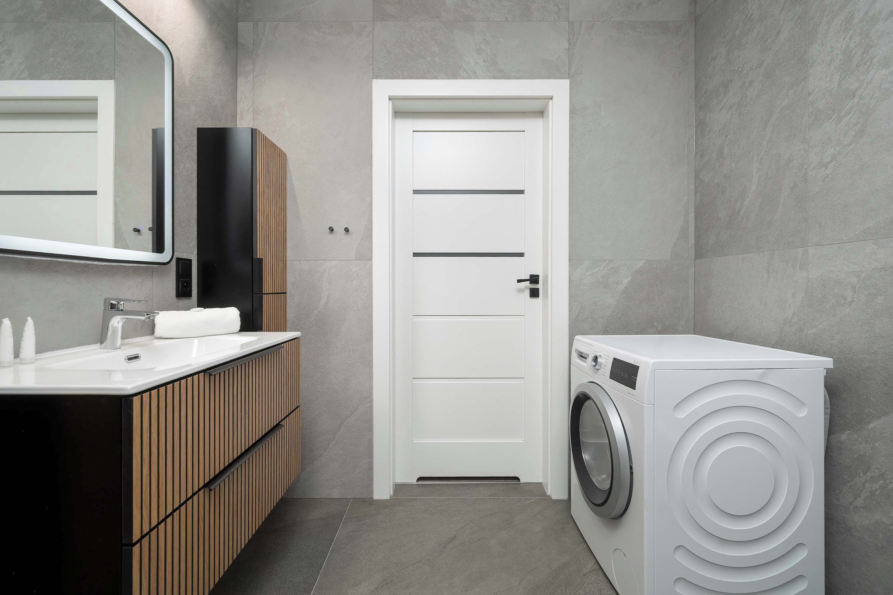 Casa de banho moderna com porta branca, lavatório com espelho e máquina de lavar roupa com abertura frontal.
