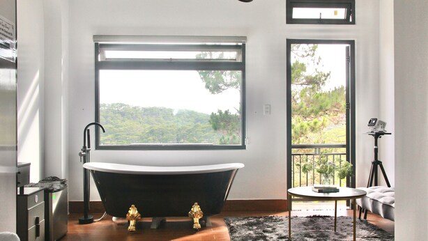 A modern bathroom with a freestanding bathtub, a large window with a view of greenery, and a small round table with a plant. - Thai Bao Chau 3  (Xuan Huong - Da Lat)