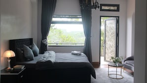 A bedroom with a bed, a nightstand, a lamp, a window with curtains, a chandelier, and a view of greenery outside. - Thai Bao Chau 3  (Xuan Huong - Da Lat)
