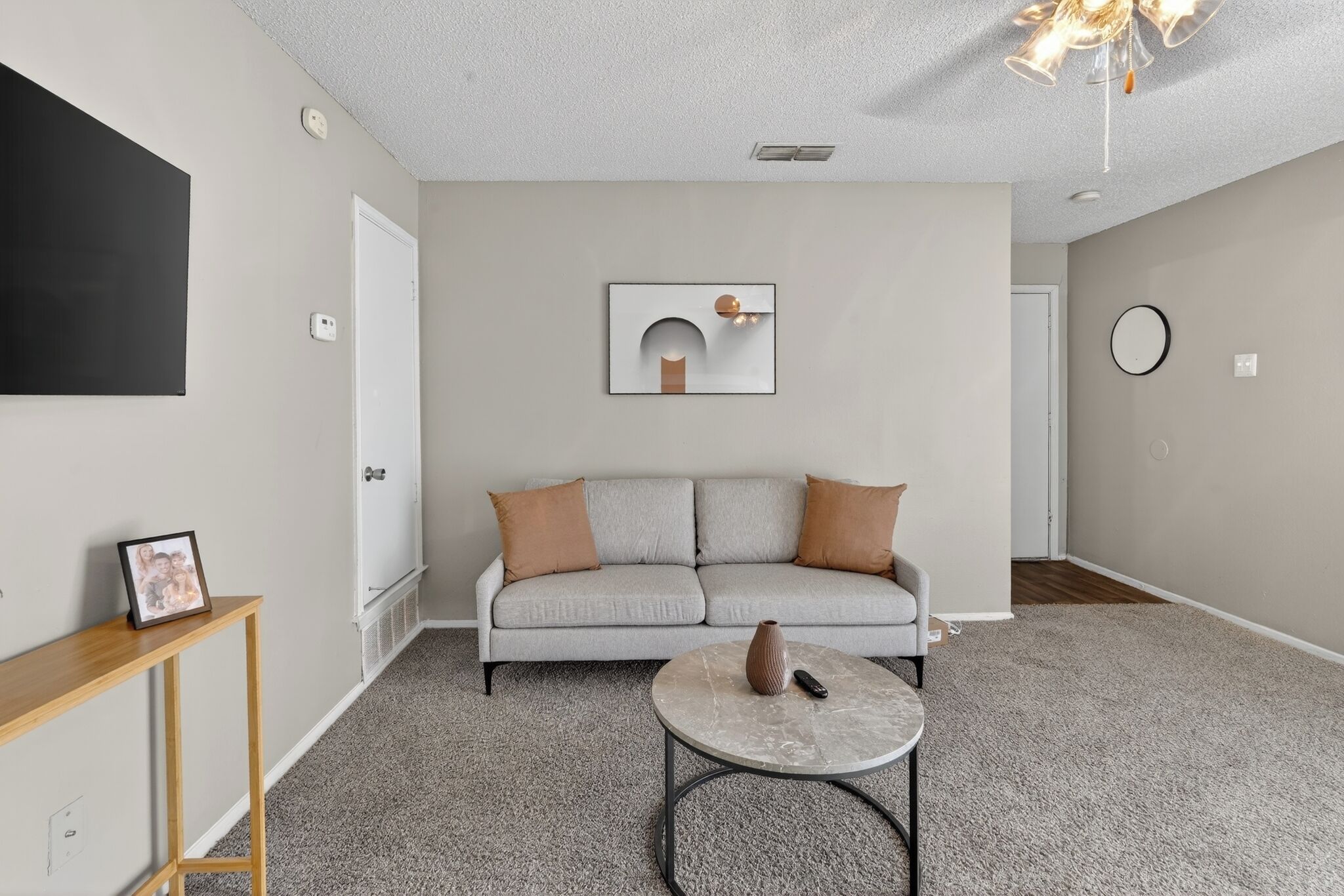 A living room with a grey sofa, a small round coffee table, a picture frame on a side table, a wall-mounted artwork, and a ceiling light fixture.
