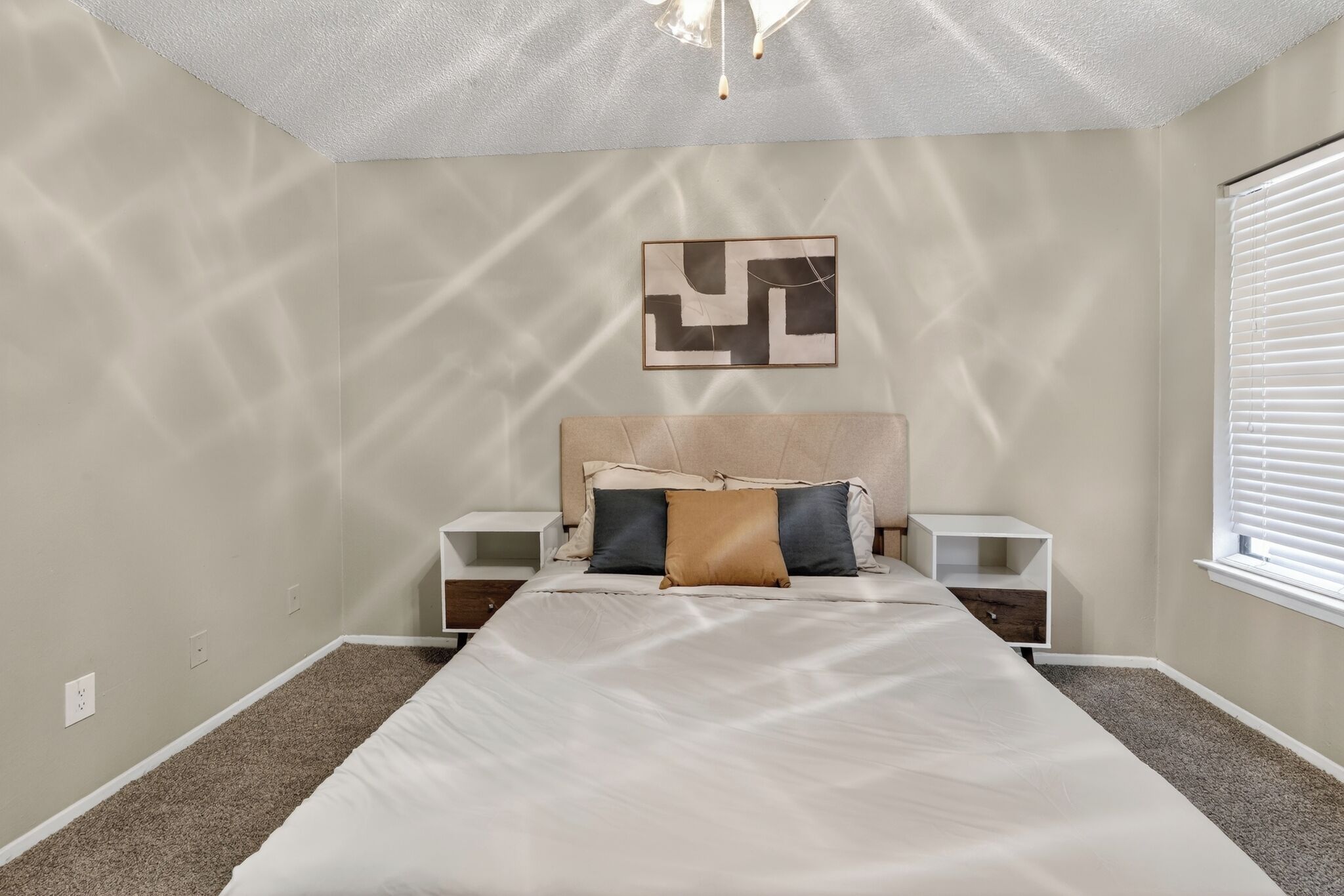Une chambre à coucher comprenant un lit, des tables de chevet, une œuvre d’art accrochée au mur et une fenêtre avec des stores.