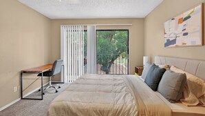 Ein Schlafzimmer mit Bett, Schreibtisch, Stuhl und einem Balkon mit Blick auf Bäume, sowie einem Bild an der Wand.