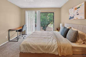 A bedroom with a bed, a desk, a chair, a balcony with a view of trees, and a painting on the wall. - Well-appointed 2BR in prime location (Lubbock)