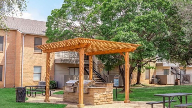 Une pergola en bois avec un barbecue, des tables de pique-nique et, au fond, un bâtiment en briques.